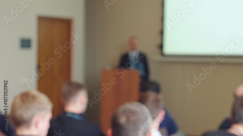 Defocused video of a speaker presenting to an audience in a meeting room. Blurred background with screen graphics, perfect as an active backdrop for titles.