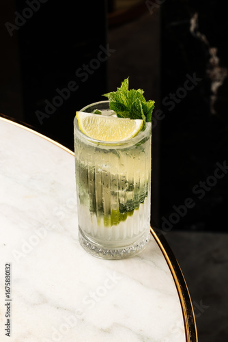 Fényképezés efreshing cocktail served in a tall glass with ice, lime wedges, and fresh mint leaves on a marble table