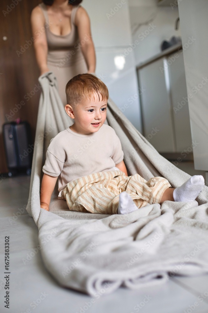 Fototapeta premium Happy child spending time at home while mother playfully pulls him on a blanket across the floor