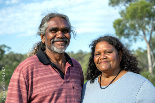An australian aboriginal couple stands together in a lush landscape, smiling brightly under a clear blue sky, enjoying the warmth of a sunny day in nature. Generative AI.