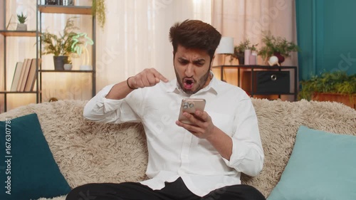 Indian man at home on sofa talking on speakerphone, gesturing intensely, arguing expressing strong emotions. Guy debating over phone call frustrated passionate reacting emotionally during conversation