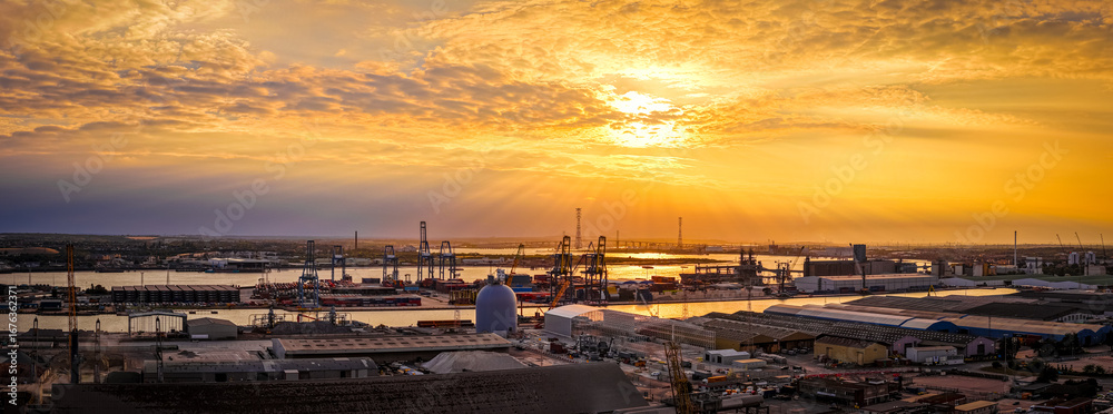 Obraz premium Golden hour drone shot of the busy Port of London, highlighting industrial activity, cranes, and shipping containers by the River Thames. Perfect for logistics, trade, and infrastructure visuals