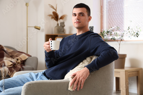 Handsome young man with cup of coffee sitting on armchair in modern living room