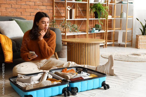 Young woman with mobile phone packing winter clothes in suitcase at home