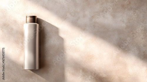 A light beige, textured surface displays a sleek, muted-tone bottle with a bronze cap, bathed in natural sunlight.