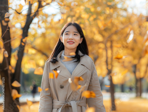 Elegant young Asian woman in trench coat walking in park with golden autumn leaves
