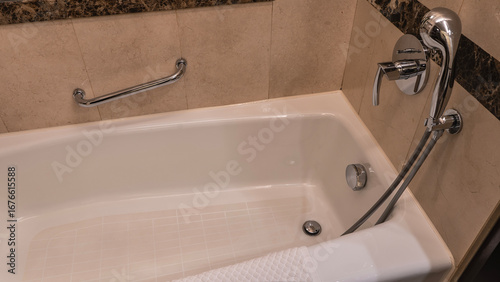 Rectangular acrylic bathtub. A fragment. Drain hole at the bottom. There is a faucet, shower, and metal handrails on the beige tiled walls. A towel on the edge.