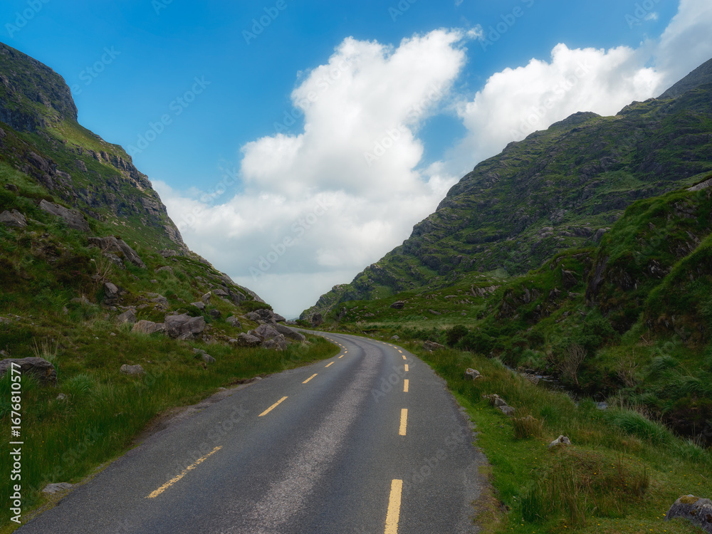 Naklejka premium A winding road cuts through a lush green valley, nestled between imposing mountains under a vibrant blue sky dotted with fluffy white clouds. The scene evokes a sense of adventure and tranquility.