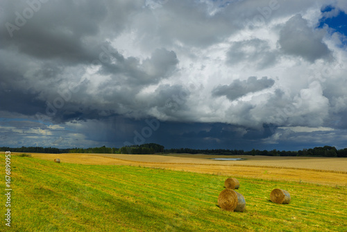 A dramatic sky with dark storm clouds looms over golden fields and green meadows dotted with hay bales.