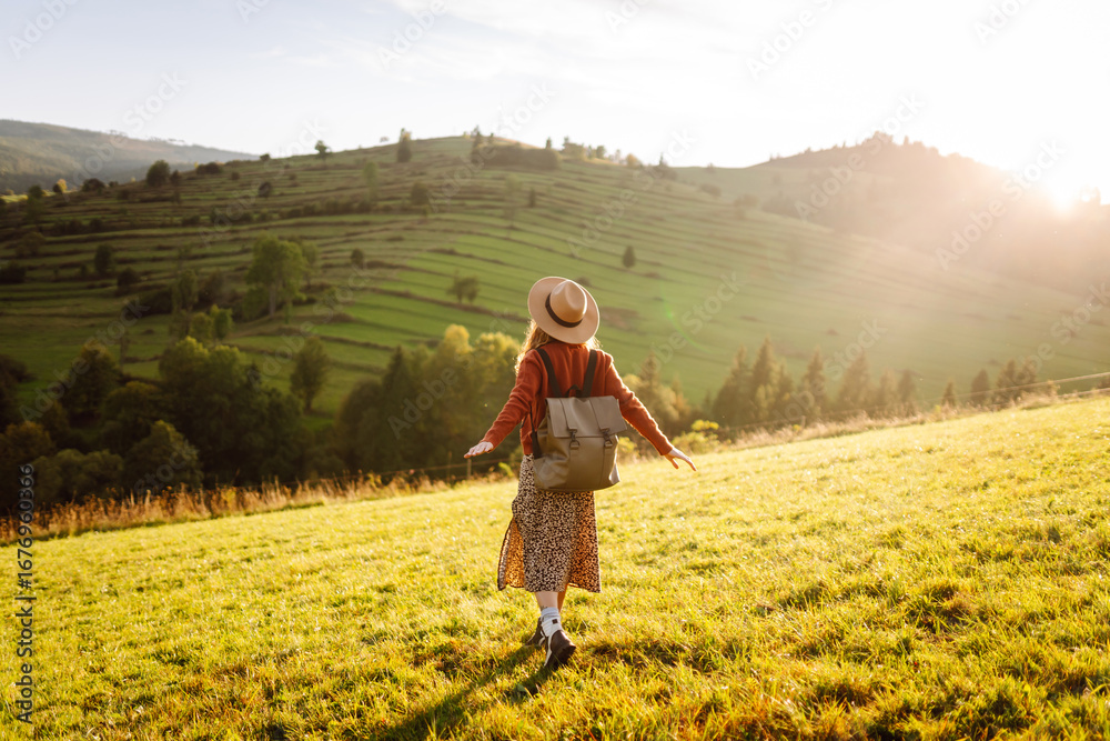 Fototapeta premium A joyful woman with a backpack and a hat walks through green hills at sunset. A traveler enjoys nature in the mountains and feels free. Active lifestyle.