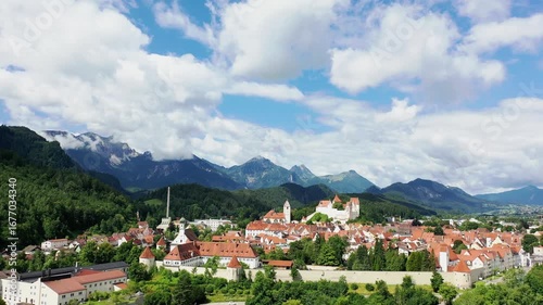 Das Foto zeigt die charmante Altstadt von Füssen mit ihren roten Dächern, liebevoll restaurierten Fassaden und dem markanten Turm der Kirche St. Mang. Im Hintergrund erhebt sich das beeindruckende Alp