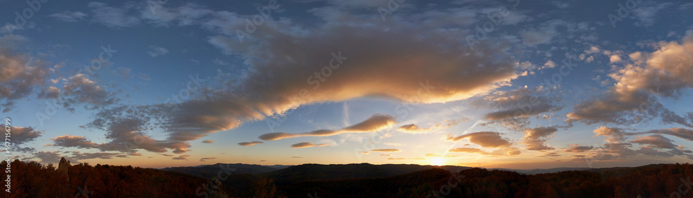 Fototapeta premium Stunning sunset fills sky with rich hues of blue and orange. Dramatic clouds with hints of pink and gold spread across sky. Silhouettes of hills in foreground add depth to breathtaking evening scene.