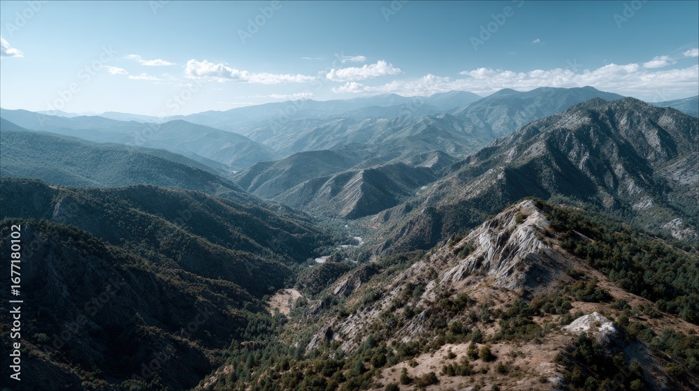 Naklejka premium A sweeping vista showcases a mountain range, with deep valleys and lush greenery, under a clear blue sky dotted with fluffy clouds.