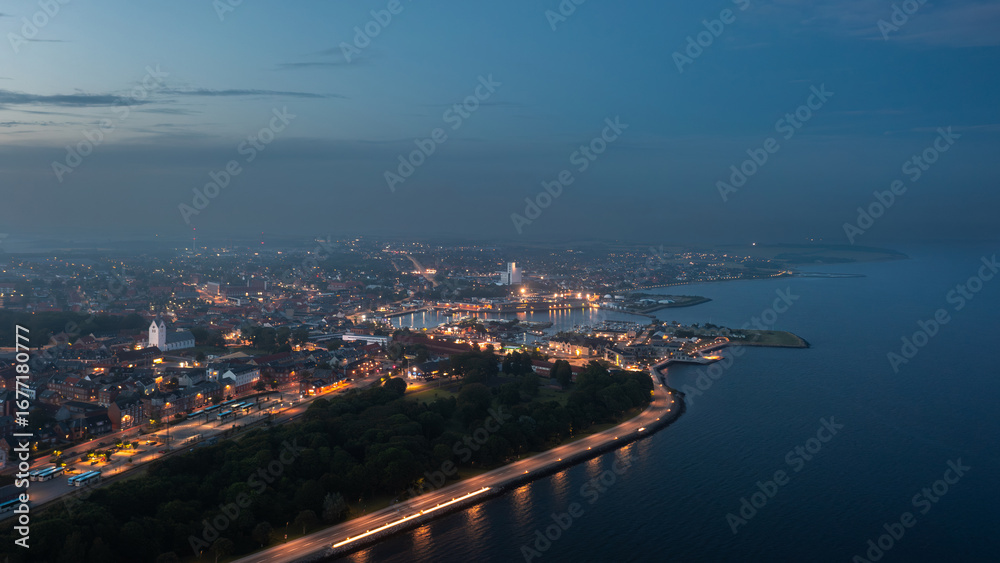 Obraz premium Aerial night view of Thisted, Denmark. Town lights, marina, Limfjord coastline, and surrounding neighborhoods create a tranquil Nordic cityscape under a twilight sky