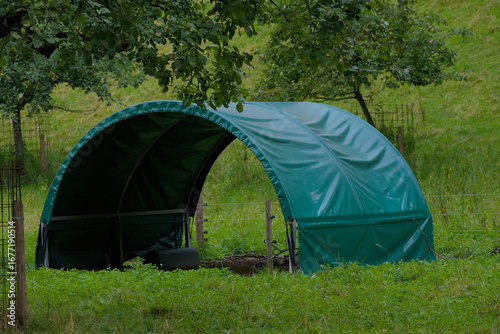 A small, green, covered shelter stands in a lush, grassy field on a cloudy day.