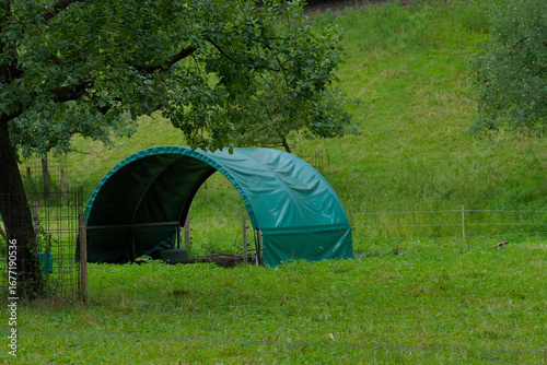 A small, green, covered shelter stands in a lush, grassy field on a cloudy day.