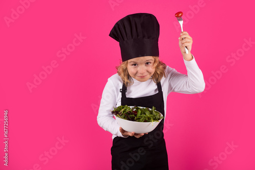 Kid cook hold plate with vegetables and fork with tomato. Chef kid preparing healthy food. Portrait of child with chef hats.