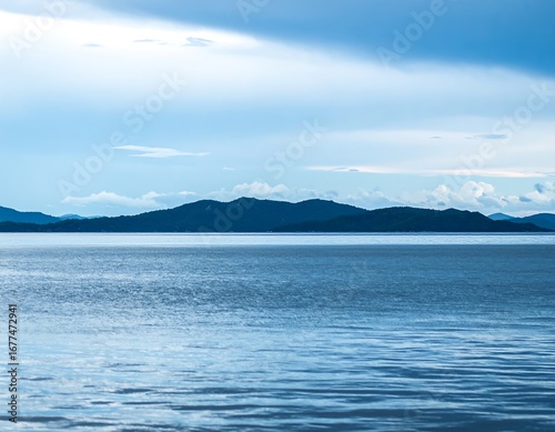 Calm, wide expanse of blue water meets a distant dark mountain range under a cloudy sky