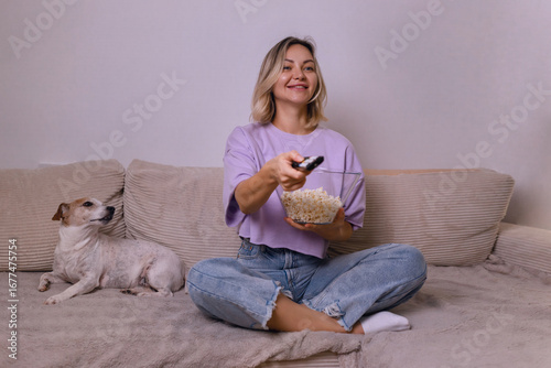 Happy young woman watching TV and smiling, holding popcorn bowl, sitting on sofa with cute dog. Cozy home evening, relaxing time with pet and favorite show.