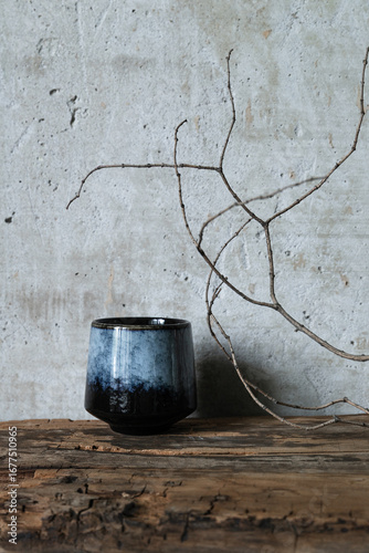 Minimalist still life with a dark ceramic vase and dry branch on rustic wood against a textured concrete wall. Wabi-sabi style composition