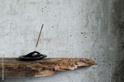 Burning incense stick on a rustic holder with ash, placed against a textured concrete wall with dry branches and old wood. Minimalist still life in wabi-sabi style.