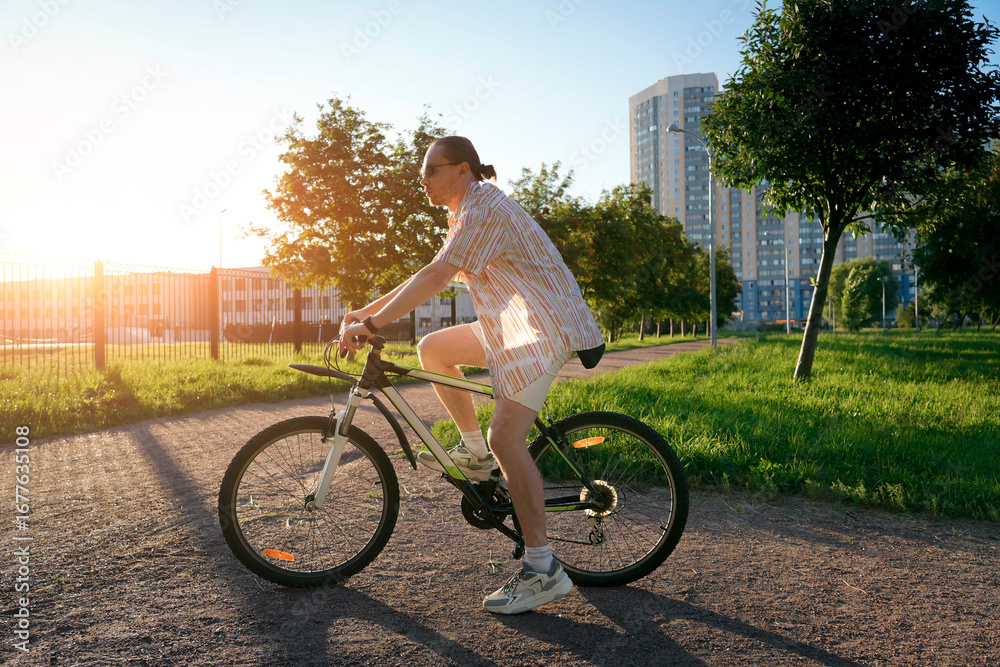 Obraz premium A man with a ponytail is riding a bicycle along a paved path in a park. The sun is setting and creating a golden hour light, casting a flare in the sky. Tall buildings are in the distance