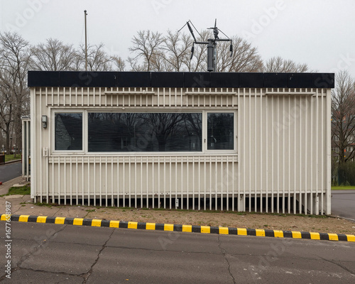 Closed guardhouse with vertical metal siding and a large front window, located by a roadside with black and yellow curb markings.