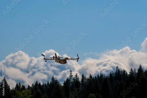 Firefighting aircraft flying low over forest with cloudy background.