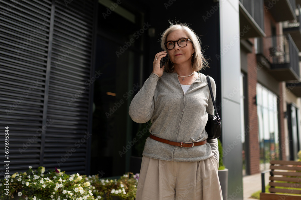 Fototapeta premium Elderly woman talking on phone while standing outside modern building in urban area