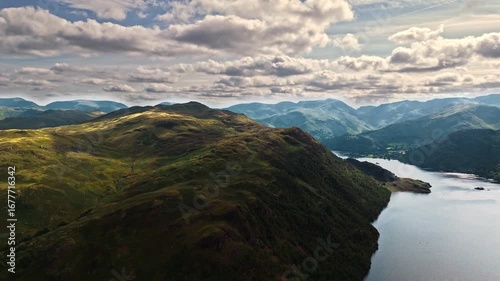 aerial drone flyover of lake ullswater inside mountain valley facing mountain covered in grass and trees in summer with blue cloudy skies