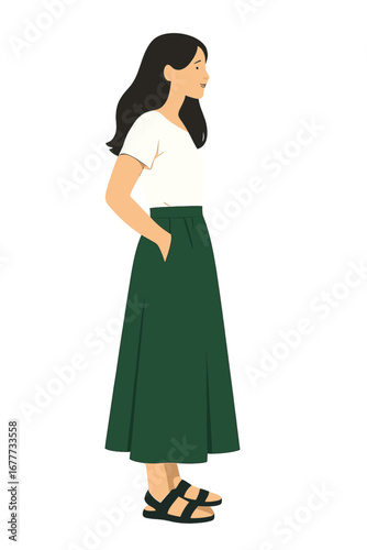 A young woman poses in a studio with a light backdrop, wearing a long green skirt, a white summer T‑shirt, and black sandals, standing sideways with her hands in her pockets