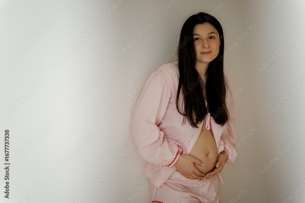 Obraz premium A young mom in a pink checkered outfit with a heart design, standing by a window. She has long dark hair and is looking thoughtfully outside, surrounded by indoor plants