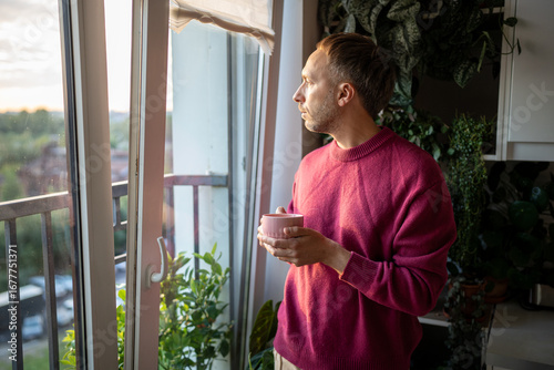 Pensive introspective man with hot tea drink in solitude looking out window in sadness deep reflection. Lonely thoughtful calm male by the window, emotional calmness, time for tea.