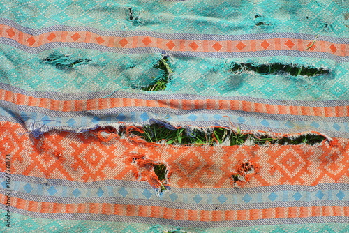 texture of old torn green with orange patterns fabric with horizontal long holes and uneven edges lies on the grass