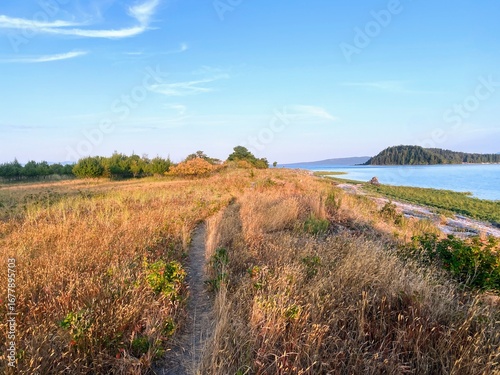 Coastal landscape on Sandy Island near Denman Island, British Columbia. Narrow dirt trail through golden grass meadow, blue ocean shoreline, forested hills, summer sky, Canadian nature, hiking, wilder