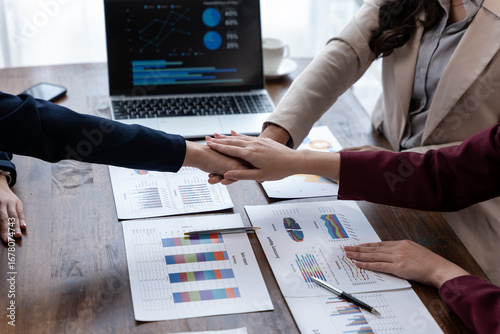 Team members stack hands over financial reports and charts on a desk, aligning on strategy during a meeting, with a laptop displaying analytics.