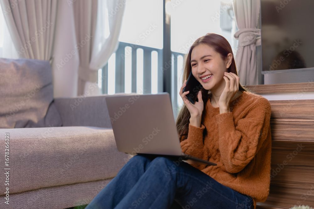 Naklejka premium Happy businesswoman working remotely from home, sitting comfortably on the floor with a laptop on her lap, talking on the phone and smiling cheerfully while enjoying her work