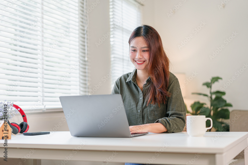 Fototapeta premium Young woman sitting at laptop smiling in bright home office with coffee and plant