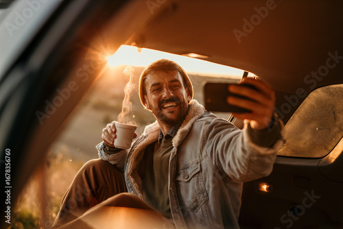 Smiling man taking selfie while enjoying hot coffee during road trip at sunset