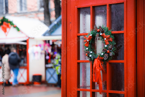 Classic red England telephone cabin with wreath on Christmas fair pedestrian market square place with foreground decorative elements and blurred background view