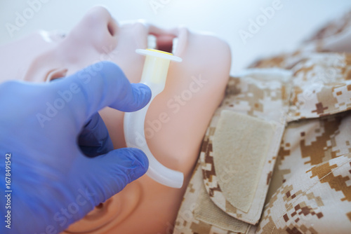 Blue-gloved hand measuring an oropharyngeal airway tube against a medical training mannequin in a camouflage uniform to check the correct size for airway management.
