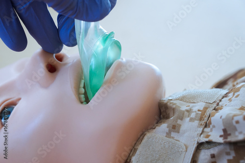 Medical training mannequin in camouflage uniform during insertion of an i-gel supraglottic airway device. The procedure illustrates airway management placement as part of a military medical simulation