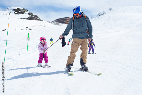 Front view of a father pulling his little daughter with his ski pole on a green slope during a nice ski day.