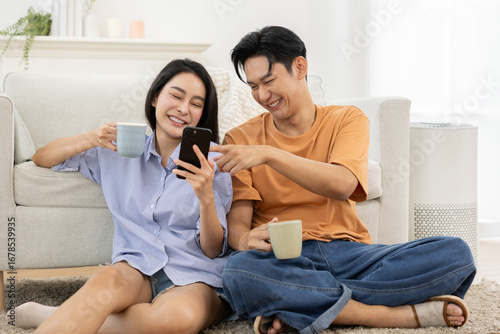 Asian Young couple sitting on floor in living room, smiling and enjoying coffee while looking at smartphone together, casual home setting with cozy atmosphere