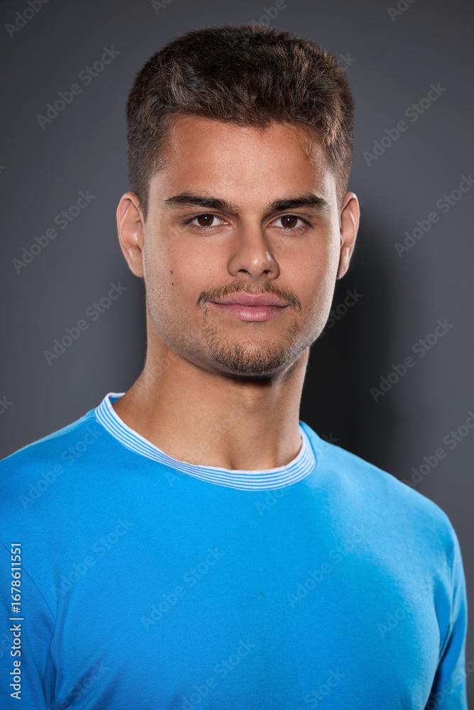 Fototapeta premium Portrait of an attractive young brunette with a short haircut and light stubble in a blue T-shirt on a gray background