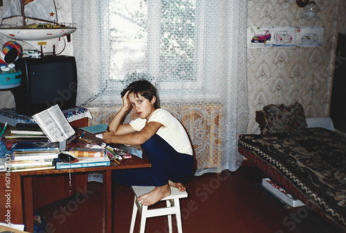 Boy does school homework sitting on stool at home table, captured in analog photography style with mid-1990s interior in Ukraine