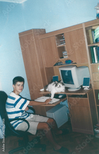 Analog photo from early 2000s Ukraine showing a teenager sitting at a home computer desk with a cat nearby, warm interior lighting