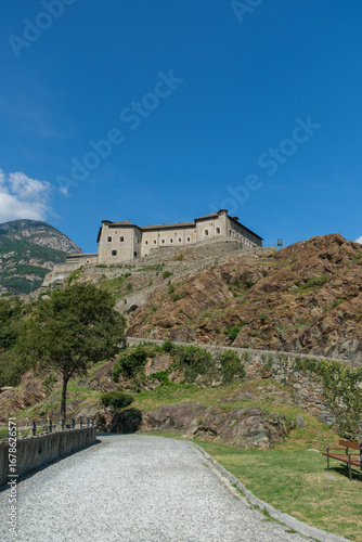 The majestic Fortress of Bard, a monumental military structure, stands imposingly on a rocky cliffside under a clear blue sky in Italy's Aosta Valley.