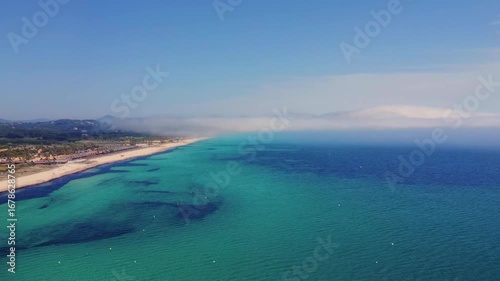 An aerial drone shot captures the vibrant scene of Pampelonne Beach in Ramatuelle, French Riviera, during a sunny summer day. The turquoise Mediterranean Sea meets the sandy shoreline, dotted with num