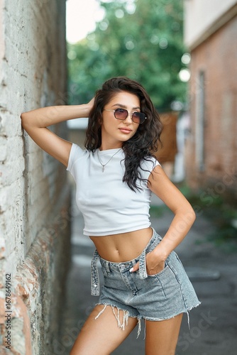 A stylish young woman in sunglasses and denim shorts poses in an urban alleyway.